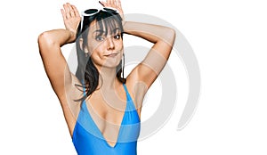 Young hispanic woman wearing swimsuit and sunglasses doing bunny ears gesture with hands palms looking cynical and skeptical