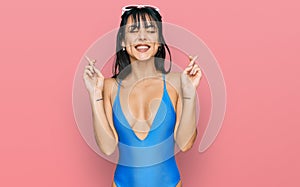 Young hispanic woman wearing swimsuit and sunglasses gesturing finger crossed smiling with hope and eyes closed