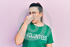 Young hispanic woman wearing volunteer t shirt smelling something stinky and disgusting, intolerable smell, holding breath with