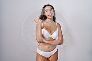 Young hispanic woman wearing white lingerie smiling with happy face looking and pointing to the side with thumb up