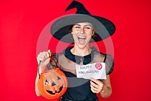 Young hispanic woman wearing witch costume holding pumpkin and happy halloween message smiling and laughing hard out loud because