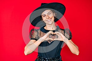 Young hispanic woman wearing witch halloween costume smiling in love showing heart symbol and shape with hands