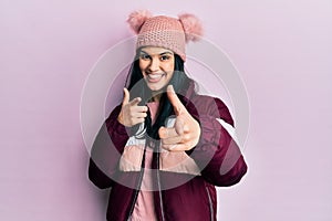 Young hispanic woman wearing wool winter sweater and cap pointing fingers to camera with happy and funny face