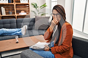 Young hispanic woman working as psychology counselor smelling something stinky and disgusting, intolerable smell, holding breath