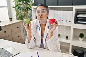 Young hispanic woman working at dietitian clinic holding green apple puffing cheeks with funny face
