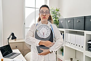 Young hispanic woman working at dietitian clinic puffing cheeks with funny face