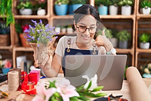 Young hispanic woman working at florist shop doing video call smiling doing phone gesture with hand and fingers like talking on