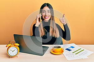 Young hispanic woman working at the office with laptop talking on the smartphone smiling happy and positive, thumb up doing