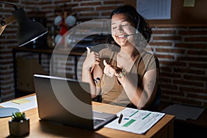 Young hispanic woman working at the office at night pointing to the back behind with hand and thumbs up, smiling confident