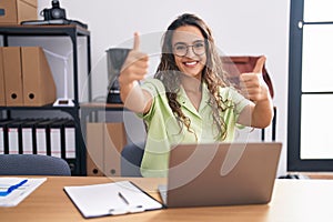 Young hispanic woman working at the office wearing glasses approving doing positive gesture with hand, thumbs up smiling and happy