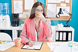 Young hispanic woman working at the office wearing glasses smelling something stinky and disgusting, intolerable smell, holding