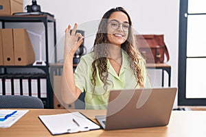 Young hispanic woman working at the office wearing glasses smiling positive doing ok sign with hand and fingers