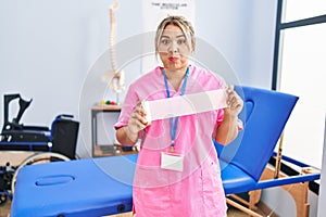 Young hispanic woman working at pain recovery clinic holding bandage puffing cheeks with funny face