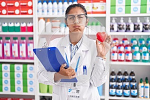 Young hispanic woman working at pharmacy drugstore holding red heart puffing cheeks with funny face