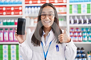 Young hispanic woman working at pharmacy drugstore showing smartphone screen smiling happy and positive, thumb up doing excellent