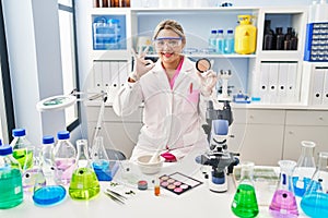 Young hispanic woman working at scientist laboratory doing make up doing ok sign with fingers, smiling friendly gesturing