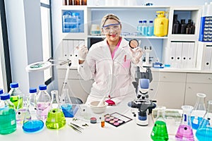 Young hispanic woman working at scientist laboratory doing make up pointing thumb up to the side smiling happy with open mouth