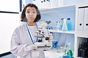 Young hispanic woman working at scientist laboratory holding microscope puffing cheeks with funny face