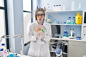 Young hispanic woman working at scientist laboratory holding money banknotes afraid and shocked with surprise and amazed