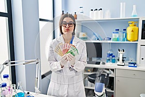 Young hispanic woman working at scientist laboratory holding money banknotes puffing cheeks with funny face