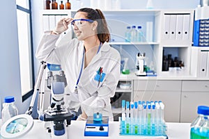 Young hispanic woman working at scientist laboratory very happy and smiling looking far away with hand over head