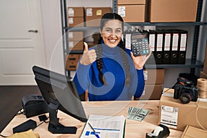 Young hispanic woman working at small business ecommerce holding calculator smiling happy and positive, thumb up doing excellent