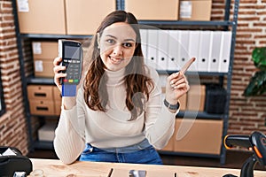 Young hispanic woman working at small business ecommerce holding dataphone smiling happy pointing with hand and finger to the side