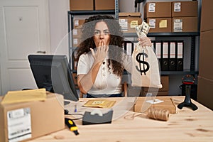Young hispanic woman working at small business ecommerce holding money bag covering mouth with hand, shocked and afraid for