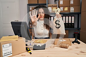 Young hispanic woman working at small business ecommerce holding money bag with open hand doing stop sign with serious and