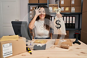 Young hispanic woman working at small business ecommerce holding money bag smiling happy doing ok sign with hand on eye looking