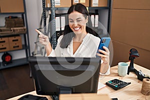 Young hispanic woman working at small business ecommerce with smartphone smiling happy pointing with hand and finger to the side