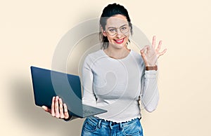 Young hispanic woman working using computer laptop doing ok sign with fingers, smiling friendly gesturing excellent symbol
