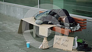 Young homeless drunk man preparing to sleep on cardboard on bench at the sidewalk