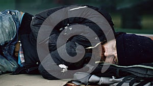 Young homeless drunk man trying to sleep on cardboard on bench at the sidewalk