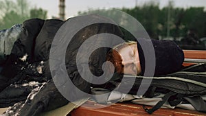 Young Homeless man trying to sleep under jacket on bench at the sidewalk