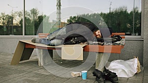 Young Homeless man trying to sleep under jacket on bench at the sidewalk