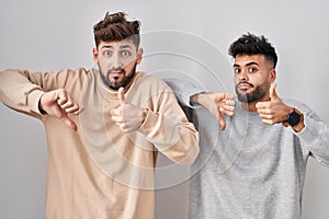Young homosexual couple standing over white background doing thumbs up and down, disagreement and agreement expression