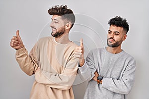 Young homosexual couple standing over white background looking proud, smiling doing thumbs up gesture to the side