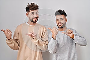 Young homosexual couple standing over white background pointing to the back behind with hand and thumbs up, smiling confident