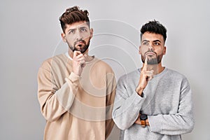 Young homosexual couple standing over white background thinking concentrated about doubt with finger on chin and looking up