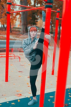 Young hooded ballerina woman warming up at playground pillars and doing splits in park at autumn