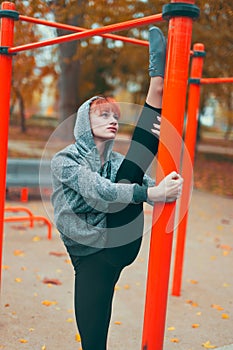 Young hooded gymnast woman warming up at playground pillars and doing splits and focusing in park at autumn