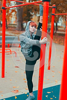 Young hooded martial artist woman warming up at playground pillars and doing splits in park at autumn