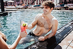 Young hot couple resting at swimpool. Sexy guy in water holding cocktails in hand and cheering with woman. Resort spa
