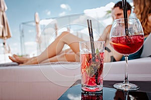 Young hot couple resting at swimpool. Two red cold tasty cocktails stand on table. Man and woman lying behind and