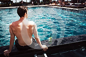 Young hot man resting at swimpool. Back view of guy sitting at water alon and look forward. Enjoy his time lonely in