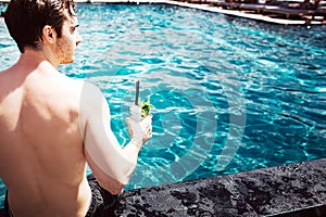Young hot man resting at swimpool. Cut back view of strong powerful guy holding cocktail in hand and enjoy spending time