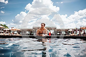 Young hot man resting at swimpool. Front view of sexy well-built guy with red cocktail lying at water and enjoy his