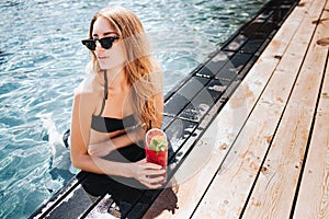 Young hot woman resting at swimpool. Girl in sexy dark bikini and sunglasses holding red cocktail and posing on camera