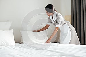 Young hotel maid setting up pillow on bed